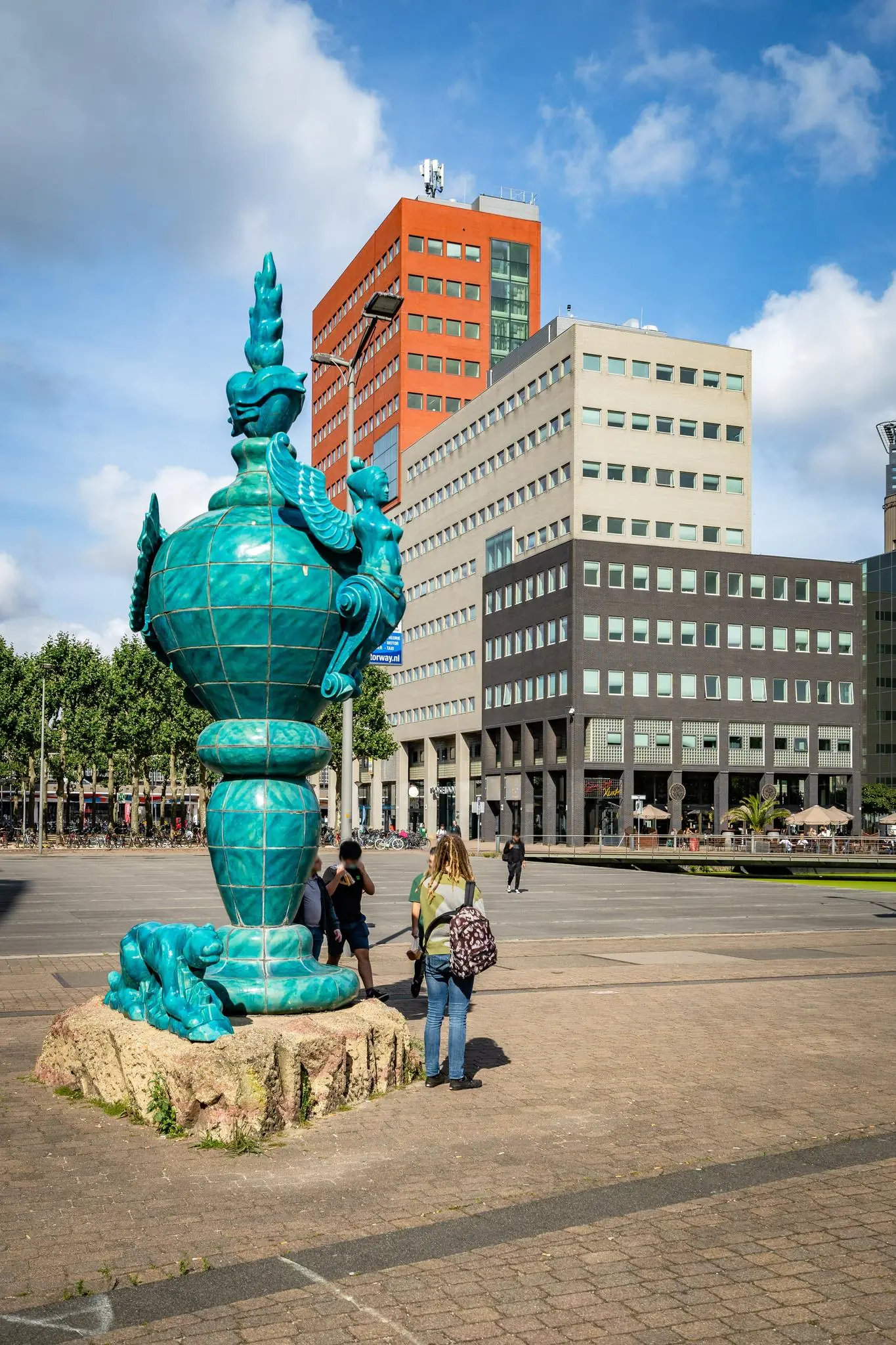 Blauwgroen keramisch kunstwerk op het Johanna Westerdijkplein in Den Haag met moderne kantoorgebouwen op de achtergrond.